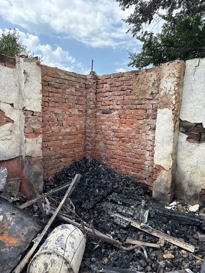 The scene depicts the fire-damaged interior corner of a structure in Nedryhailiv, Ukraine. The two visible walls are constructed of red brick, partially covered by deteriorated, cracked, and peeling light-colored stucco or plaster, exposing the underlying brickwork. The top edges of the walls are irregular, indicating the absence of a roof. A single metal rod protrudes vertically from the top of the corner joint. The ground within the corner is covered by a dense layer of black, charred debris, consistent with burnt organic materials. Embedded within this debris are multiple heat-distorted metal objects, including a prominent, light-colored cylindrical tank or drum in the lower-left foreground, lying horizontally and appearing scorched and discolored. Other indeterminate metal fragments and charred wooden remnants are interspersed throughout the debris field. The upper background reveals a clear blue sky with white clouds and the green foliage of trees on both sides, visible above the remaining wall structure. No people or animals are present.