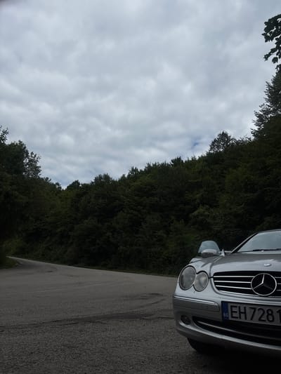 In Shipkovo, Bulgaria, a silver Mercedes-Benz CLK convertible is parked on the side of a winding asphalt road. The car's front is partially visible, including its headlights, grille with the Mercedes-Benz logo, and a license plate reading "EH 7281 BG". The road curves to the left and disappears into the distance, bordered by a dense, dark green forest. The sky above is overcast with gray clouds. The image appears to have been taken from a low angle, emphasizing the car and the surrounding natural environment.
