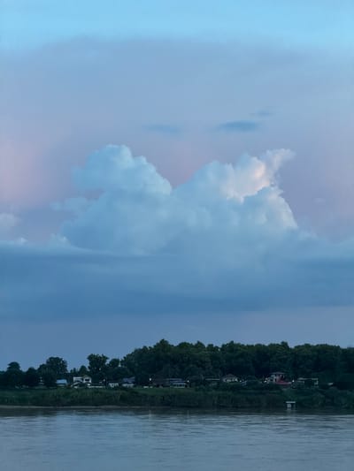 The image shows a dusky sky over the Mekong River in Vientiane Capital, Laos. The sky transitions from a pale blue at the top to a deeper blue near the horizon, with soft pink and white hues reflecting off the clouds. A large, prominent cloud formation dominates the upper half of the sky, appearing sculpted and luminous against the twilight. Below the sky, a dense line of trees forms a dark green canopy along the riverbank. Scattered among the trees are several buildings, some appearing to be houses with lights on, suggesting a small village or residential area. The Mekong River occupies the bottom portion of the image, its surface calm and reflecting the muted colors of the sky and the surrounding landscape. The overall scene conveys a peaceful and serene evening atmosphere.