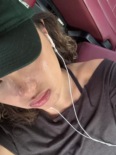 The image shows a close-up of a person's face and chest. They are wearing a green baseball cap, a gray t-shirt, and white earbuds. They are sitting in a vehicle with a red seat. The person's eyes are closed and their lips are slightly parted. They are likely listening to music or a podcast. The image was taken in the city of 信義里, Taiwan. The location is determined by the coordinates (latitude: 22.785, longitude: 120.297).