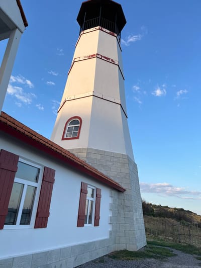 The image shows a white lighthouse with a red-trimmed lantern room, a white building with red-shuttered windows and a red-tiled roof, and a gentle slope covered in dry grass and a rustic wooden fence in the background. The sky is blue with scattered white clouds. The scene is static and depicts the exterior of the buildings and the surrounding landscape. The location is identified as the city of Merzhanovo, Russia.