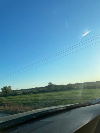 The image depicts a view from inside a vehicle, likely a car, moving through the Xanthi Municipal Unit, Greece. The sky is clear blue with a few wispy clouds and power lines cutting across the field of view. To the right of the sky, the landscape features green fields and trees, giving way to rolling hills in the distance. Part of the car's interior, including a window frame and dashboard, is visible on the right side of the image. The scene appears to be taken during daylight.