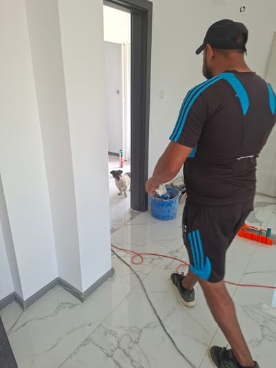 A man wearing a black baseball cap, black t-shirt with blue stripes, and black shorts with blue stripes is walking through a doorway. He is carrying a blue bucket filled with materials. In the background, a small dog with tan and black markings is looking towards the man. The scene appears to be an interior of a building under construction, with white walls and marble-patterned tiled floors. Tools and building materials are visible in the background. The location is identified as Dolna Malina, Bulgaria.