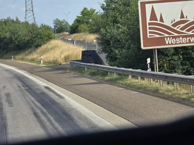 The image shows a highway scene in Windhagen, Germany. A brown sign with white stylized hills and the text "Westerw" is visible on the right side of the road. The highway itself has tire marks on the asphalt. A metal guardrail runs along the side of the road, separating it from a grassy embankment with some trees and vegetation. In the background, there is a large power pylon, a fence, and more trees. A dark, angular object, possibly a speed camera or surveillance equipment, is positioned behind the guardrail. The sky is clear and blue.