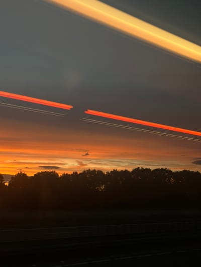 The image captures a dynamic sunset scene with streaks of light, likely from motion blur due to a moving vehicle. The sky is a vibrant gradient of orange and yellow, transitioning into a darker gray at the top. Silhouetted against the colorful sky is a dense line of trees, forming a natural horizon. Below the trees, the foreground is dark, suggesting a road or landscape obscured by the fading light. The specific location in Bubenreuth, Germany, is not visually identifiable from the provided image alone, as there are no distinguishing landmarks or features. The image appears to be taken during twilight or dusk.