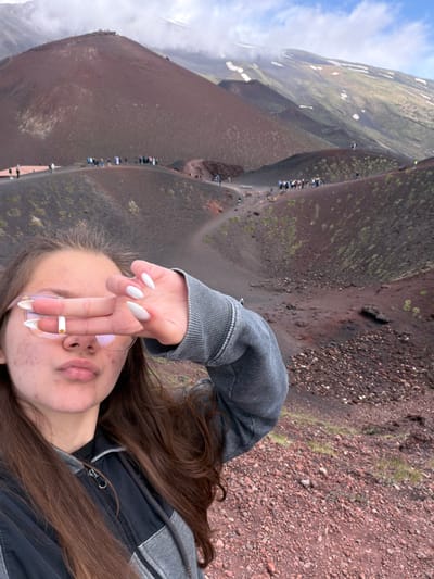 A selfie-style, rotated shot depicts a person with light skin, brown hair, and a grey and black jacket, holding a white, ring-like object, possibly a cigarette, between their fingers. The location is suggestive of Mount Etna near Nicolosi, Italy, given the volcanic terrain of red-brown rock, sparse vegetation, and a visible path with groups of people in the distance. The sky is partially obscured by a mountain on the left.