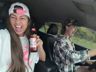 This media file depicts two women inside a car. The woman on the left is wearing a black and white plaid shirt and appears to be laughing. The woman on the right is wearing a white t-shirt with pink lettering and a white baseball cap with a pink band. She is holding a bottle of Budweiser beer and has her mouth open, appearing to be laughing or shouting. Both women are positioned upside down in the frame, suggesting the photo was taken from an unusual angle or has been rotated.

The setting appears to be daytime, with greenery visible through the car windows, indicating an outdoor environment. The overall mood seems to be lighthearted and fun, with both women displaying expressions of joy. The visible text on the Budweiser bottle reads "Budweiser." The t-shirt of the woman on the right also has pink lettering, though it is partially obscured and difficult to read. The background elements suggest a rural or suburban landscape.
