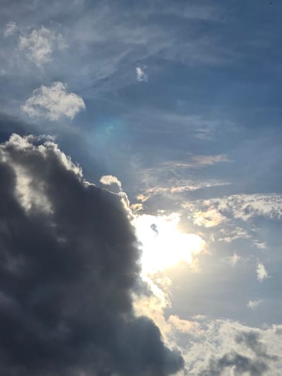 The image shows a dramatic sky with a large, dark cloud dominating the left side. The sun is peeking out from behind this cloud, creating a bright, almost blinding light that illuminates the edges of the cloud and a portion of the sky. Wisps of lighter clouds are scattered across the blue sky, some catching the sunlight and appearing golden. The sky suggests a dynamic weather event, possibly the transition from a storm or the approach of one. The location is identified as Kostelec na Hané, Czechia. There are no people, specific subjects, or discernible objects other than the clouds and the sun. The scene is purely atmospheric and natural, conveying a sense of awe and the power of weather.
