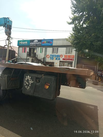 This image depicts a street scene in Novopavlovka, Kyrgyzstan, on June 19, 2026, at 12:09 PM. A truck with a flatbed and an attached crane is parked on the right side of the road. The truck's rear section, including a black fuel tank with a stylized lion emblem and an acorn sticker, is prominently visible in the foreground. Several people are standing and observing the scene near a building on the right side of the frame. The building features multiple signs, including "Mbank," "БЫЛДАМ КАРГО" (BOLD CARGO), "МАДОШКИ" (MADOSHki), and "TECHNO TOЧKA" (TECHNO POINT) with "РАССРОЧКА" (INSTALLMENT) below it. The environment is urban, with a paved road and power lines overhead. A large tree with dense green foliage partially obscures the sky and the building on the right. The weather appears sunny, casting shadows on the road.