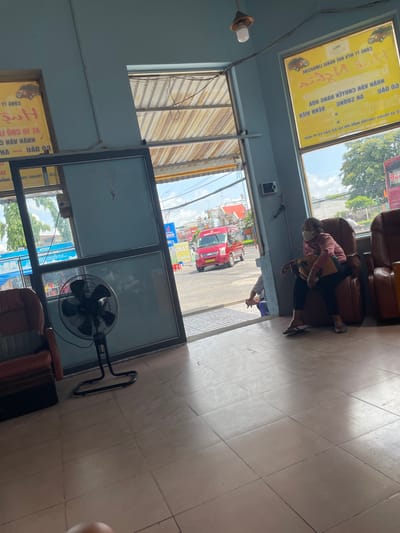 The image depicts the interior of what appears to be a bus station or waiting area in Gò Dầu, Vietnam. A woman wearing a face mask and a pink long-sleeved shirt is seated in a reclining chair, looking to her left. She is carrying a bag on her shoulder. To her left, a large oscillating fan is positioned on the tiled floor. Through a large window or opening, a red minibus is visible on a street, along with trees and buildings in the background. Yellow signs with text in Vietnamese are displayed on the windows. Another person is partially visible sitting on a blue stool near the opening, facing away from the camera. The overall environment suggests a functional waiting area with typical amenities for travelers.