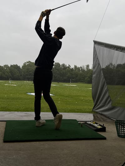 A young male golfer in black clothing, including a black hoodie, black pants, and white sneakers, is swinging a golf club at a driving range in South Ribble, United Kingdom. The golfer is facing away from the viewer and towards a large grey net, which is likely there to stop errant golf balls. The golfer is in mid-swing and has his right leg extended, while his left leg is bent. There are white golf balls scattered across the green driving range and white golf balls inside a black tray to the right of the golfer. The location is identifiable by the coordinates provided: 53.771, -2.57. The sky is overcast with a slight drizzle. The ground is wet. The driving range is near a wooded area. The golfer is alone. The scene is serene and tranquil. It is likely a weekday morning or afternoon, as there is no one else present on the driving range.