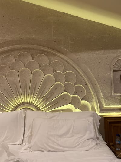 A high-angle, rotated view captures a bed in a room, likely in Ürgüp, Turkey, given the distinct architectural details. The headboard is elaborately designed, featuring a large, daisy-like carved stone pattern. The bed is covered in crisp white linens. A portion of a wooden headboard or side table is visible behind the bed. A water bottle is positioned atop the wooden structure. The image is well-lit, highlighting the textures of the stone carving and the fabric of the bed linens.