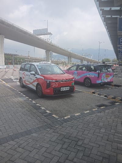 This image depicts an outdoor urban scene in Tung Chung, China, under an overcast sky. In the immediate foreground, a pedestrian area paved with grey and dark grey interlocking bricks in a herringbone pattern abuts an asphalt surface. A black and yellow striped curb marks the transition, and a storm drain grate is visible in the asphalt.

Two minivan-style vehicles are parked on the asphalt. The prominent vehicle in the foreground is a white and red taxi displaying Hong Kong license plate ZY 1650 on its front. It features a "TAXI" sign on its roof and "TAXI" lettering on its side, alongside an "S" logo. The vehicle has a red front bumper. Partially obscured behind and to the right of this vehicle is a second minivan-style vehicle, colored pink and light blue, with "ETIX" branding visible on its side and rear. Both vehicles appear stationary.

In the midground and background, a large, elevated concrete bridge or highway structure with multiple lanes and numerous concrete support pillars spans the view. To the far right, a section of a building with reflective glass panels and vertical alphanumeric markings, including '5' and 'P', is visible. Distant mountains are discernible on the horizon beneath the overcast sky. Urban infrastructure such as streetlights and orange traffic cones are also present in the background. No individuals are visible in the scene.