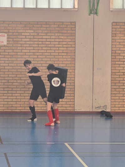 Two individuals are positioned on a blue indoor court with white lines. One person is wearing black shorts, a black shirt, and black and white shoes. The other is wearing black shorts, a black shirt with a circular logo, and red socks. They are indoors, possibly in a gymnasium, as evidenced by the brick wall, the basketball hoop, and the presence of athletic shoes placed away from the main activity. The location can be inferred as Leers, France, given the information available in the image.