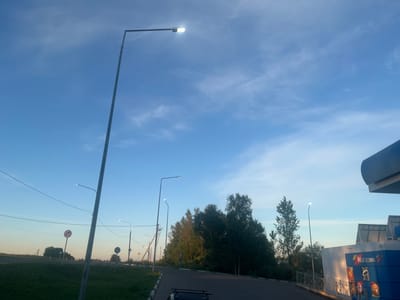 The image captures an outdoor scene in Tutaev, Russia, likely during twilight due to the blue sky with wispy clouds and illuminated streetlights. A grey metal lamppost stands prominently on the left side of the frame, its light emitting a bright glare. Further down a paved road, another lamppost is visible, also illuminated. To the right, a structure with blue paneling and the Russian word "МАГАЗИН" (store) is partially visible, suggesting it might be part of a gas station or a convenience store. Several trees with green foliage line the right side of the road. Road signs, including a circular sign with a red border and an arrow, and a triangular sign, are present in the grassy median. The overall environment appears to be a roadside setting, possibly on the outskirts of town. No people are clearly discernible in the image.