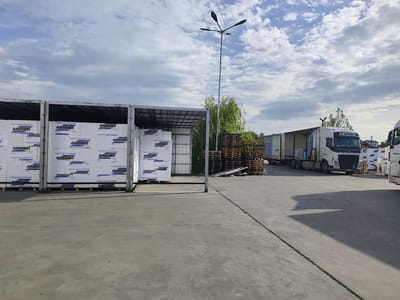 Here is a forensic description of the image:

**Location:** An outdoor industrial area in Grigorevo, Bulgaria, identified by the signage on the materials.

**Scene:** The scene displays a warehouse or storage yard filled with construction or insulation materials, identifiable by the "ANGRO" logo and descriptions (in Bulgarian) related to energy-saving thermal insulation on the packaging. These materials are stacked on pallets within open-sided storage units.

Several stacks of empty wooden pallets are visible in the background. A large white semi-truck is parked in the distance, and a second one, only partially visible, is closer to the foreground.

**Objects:**
* Palletized stacks of materials labeled "ANGRO"
* Open-sided storage units
* Wooden pallets
* Two semi-trucks
* A tall, modern street light
* Concrete pavement with expansion joints

**Environment:** Overcast sky with patches of blue, suggesting daylight conditions. The area appears to be a commercial or industrial zone.