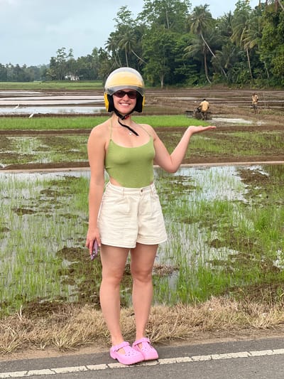 A young woman stands in front of a flooded rice paddy in Kodagoda, Sri Lanka. She is wearing a lime green crochet halter top, off-white denim shorts, a yellow motorcycle helmet, dark sunglasses, and pink clogs. She holds a purple smartphone in her left hand and her right arm is extended, palm up, in a gesture of offering. In the background, two men are working in the paddy; one is operating a tiller and the other is standing. Several white egrets are scattered throughout the field. The sky is overcast, and the surrounding area is lush with palm trees and other vegetation.