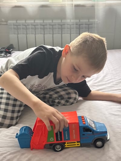 A boy is sitting on a bed and playing with a toy truck. The truck is blue and red and has labels in Russian. The boy is wearing a white and black shirt and checkered pants. The bed is covered in a white sheet. A window with a grid pattern is in the background. There is no definitive evidence to confirm the location as Fedorino, Russia.