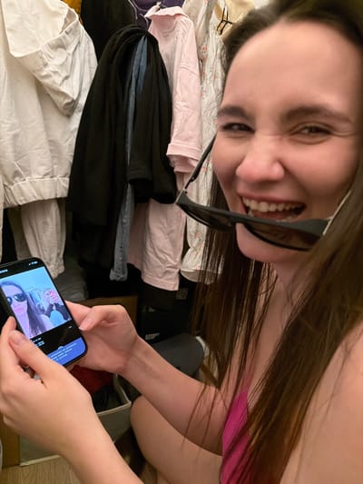 A woman with long, dark hair smiles at the camera, holding sunglasses in her mouth. She's wearing a pink top and is near a pile of clothes, including white, pink, and black items. Her hands are holding a smartphone displaying a selfie of two women with drinks. The photo's angle is tilted, making the background appear sideways.