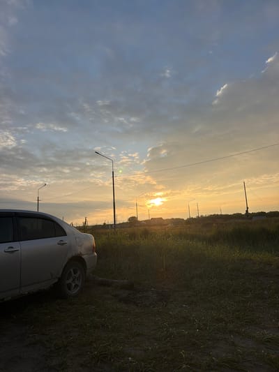 The image depicts a rural or semi-rural outdoor scene at sunset, possibly in Aniva, Russia. In the foreground to the left, a silver sedan with visible rust and damage is parked on a dirt path surrounded by tall grass. The car's rear tire is visible. The path extends into a field of green grass which is lit by the setting sun. Several lampposts are scattered throughout the scene, with power lines running between them. In the background, a line of trees and some indistinct structures, possibly houses, are visible. The sky is filled with dramatic clouds, tinged with orange and yellow from the setting sun, which is partially obscured by clouds and appears to be low on the horizon. The overall impression is one of a quiet, somewhat neglected, rural landscape at the end of the day.