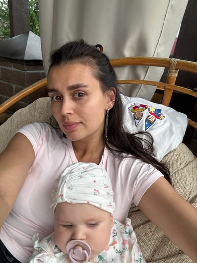 A medium shot shows a woman and a baby sitting on a chair. The woman has long dark hair and is wearing a pink t-shirt. She is looking at the camera. The baby is wearing a white hat with a floral pattern and is holding a pacifier. The baby is looking down. The background is blurred but appears to be an outdoor setting with trees and a canopy. The image was taken in the city of Sylva, Russia.