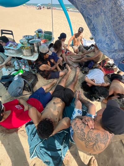 A group of young adults are gathered under a canopy on a sandy beach, with the Mediterranean Sea visible in the background. The people, mostly men, are dressed in casual beachwear and appear to be relaxing, some lying down while others are seated. They are clustered around a portable table laden with food and drinks, suggesting a picnic or outdoor meal. The overall atmosphere is informal and leisurely. The scene is consistent with a typical summer day at the beach in Gavà, Spain, a coastal town known for its beaches and recreational activities.