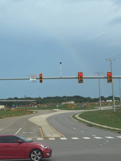 The image shows a street scene with a red car in the foreground. There are traffic lights and signs visible. In the background, there is construction activity. The location is Murfreesboro, United States. The sky is overcast. The traffic lights are red, indicating that traffic should stop. The signs indicate a left turn signal. The road has a pedestrian crossing.