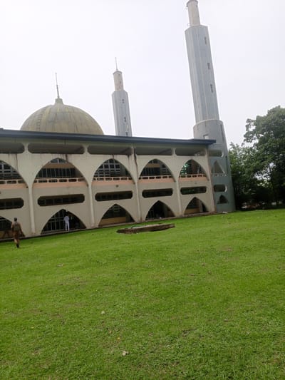 This media file shows the exterior of a mosque in Ife, Nigeria. The mosque features a large dome and two tall, slender minarets. The building's facade is characterized by a series of arched openings and a patterned concrete design. A well-maintained green lawn surrounds the mosque, with a few scattered leaves.

In the foreground, a person is walking away from the camera across the grass, wearing a brown outfit and a backpack. Further back, near the entrance of the mosque, another individual dressed in white is visible. The overall scene suggests a quiet, possibly daytime setting, with an overcast sky, indicating a cloudy or possibly rainy weather. The architecture of the mosque, with its prominent dome and minarets, is a key feature, and the surrounding lush greenery hints at a tropical or sub-tropical location. No visible text is present in the image.