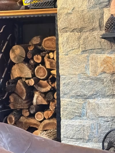 The image shows a wood-burning fireplace with a stack of firewood inside. The fireplace is built with stone masonry. The firewood consists of various sizes and shapes of logs. The grate is visible beneath the wood. The image doesn't contain any information about its location in Кирково, Bulgaria.