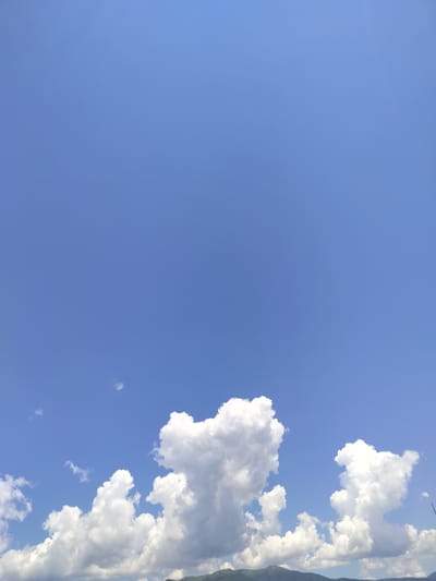 The image captures a bright, clear blue sky with a few fluffy white clouds scattered across the lower portion of the frame. In the distance, a range of hazy green mountains is visible along the horizon. The scene appears to be an outdoor landscape, likely a rural or mountainous area, given the prominent natural elements. The lighting suggests it is daytime, with sunlight illuminating the clouds and the sky. The overall impression is one of a serene and open natural environment. There are no people, specific buildings, or objects that would indicate a particular urban setting or specific actions taking place. The location is identified as Gorno Sahrane, Bulgaria.