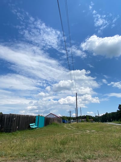 This is a medium-shot, eye-level, outdoor, daytime, vertical, full-color photograph taken in the city of Znamensk, Russia. The image depicts a rural scene with a wooden fence in the foreground, a bright teal-colored gate and bench within a grassy area. Beyond the fence, several power lines stretch diagonally across the bright blue sky, which is dotted with white cumulus clouds. A few wooden utility poles are visible, supporting the lines. Further in the background, there are residential buildings with red roofs and a dense forest line the right side of the frame. A dirt road with tire tracks curves through the grassy landscape. The overall impression is one of a quiet, rural setting on a sunny day.