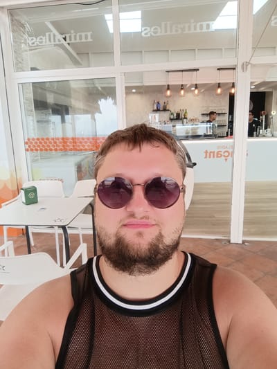 A man with a beard and sunglasses is taking a selfie. He is wearing a black mesh tank top with white trim. He is sitting at a table outside of a cafe with a view of the interior through a large glass window. In the background, there is a bar with bottles of alcohol and two bartenders. The cafe appears to be called "Milles". The interior of the cafe has wooden floors and light-colored walls. Outside, there are white tables and chairs. The man is in the city of Águilas, Spain.