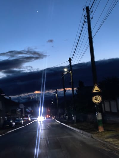The image depicts a street scene in Resilovo, Bulgaria at dusk. The road stretches into the distance, illuminated by car headlights and streetlights. Several cars are parked on the left side of the road. Overhead power lines run along the right side of the street. A traffic sign indicating a speed limit of 40 km/h is visible on the right. The sky is a mix of blue and dark clouds. A sliver of the moon is visible in the upper center of the image.