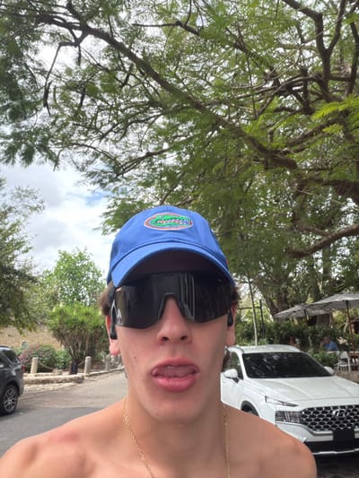 A young male individual is depicted in a medium close-up, positioned centrally and occupying the lower half of the frame, outdoors in La Romana, Dominican Republic. He wears a blue baseball cap with an embroidered "Florida Gators" logo, large dark wrap-around sunglasses, and black wireless earbuds in both ears. A gold-colored chain is visible around his neck. He is shirtless, exposing his chest and shoulders, with a minor reddish mark discernible on his left shoulder. His mouth is open, with his tongue extended slightly. The background features a vibrant tropical environment under a partly cloudy sky. Numerous large trees with dense green foliage and intricate branch structures dominate the upper two-thirds of the image. To the left, a dark-colored SUV is partially visible, parked adjacent to a paved pathway marked by light-colored stone bollards and low-lying vegetation. To the right, a white Hyundai Santa Fe SUV is parked in front of an outdoor dining area. This area includes two striped market umbrellas covering tables, with one partially visible individual seated beneath an umbrella.