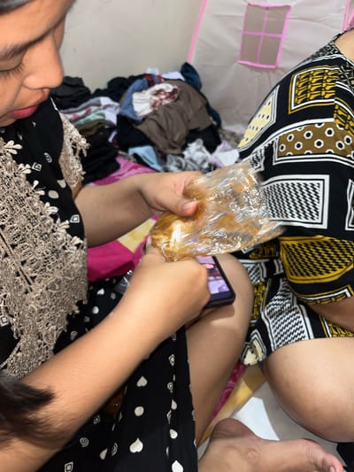 The image depicts a casual indoor scene with at least two individuals in Paweden, Indonesia. In the foreground, a person is captured from a top-down angle, wearing a black garment patterned with white polka dots, hearts, and geometric shapes, featuring a beige lace overlay. This person is actively eating from a clear plastic bag containing a food item, possibly a pastry or bun covered in a brownish sauce or glaze. Concurrently, their other hand holds a smartphone, displaying what appears to be a video or show on its screen.

Another individual is partially visible in the upper portion of the frame, dressed in a black and yellow/gold patterned garment, with their legs and bare foot in a relaxed, seated or reclining posture. The background reveals a light-colored floor with a pile of various clothes scattered informally. To the far left, a light pink and white structure, possibly a play tent or fabric enclosure, is visible. The scene conveys a relaxed and informal atmosphere, with no discernible text present.
