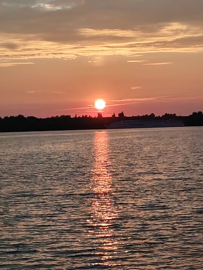 This is a landscape photograph taken at sunset over a body of water, likely a river, near the city of Selivanovo, Russia. The sun is a bright orb, partially obscured by a thin layer of clouds, casting a warm, orange glow across the sky. A reflection of the sun shimmers on the water's surface, creating a path of light that stretches towards the viewer. In the background, a dark silhouette of trees forms a treeline along the far bank. A large, multi-decked boat, possibly a river cruise ship, is visible in the distance, positioned just above the treeline. The water itself is calm, with gentle ripples disturbing its surface. The overall impression is one of tranquility and natural beauty.