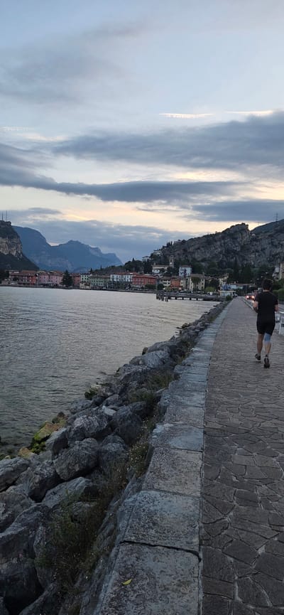 The image depicts a scenic view of the waterfront promenade in Torbole, Italy, during twilight. A lone figure, a male with dark hair wearing a black t-shirt and shorts, is jogging away from the camera on a paved walkway. The walkway is lined with large, grey rocks and overlooks a body of water, likely Lake Garda. In the background, a picturesque town with colorful buildings is nestled against a backdrop of dramatic, mountainous terrain under a cloudy sky. The overall atmosphere is serene and hints at the end of a day with subdued lighting.