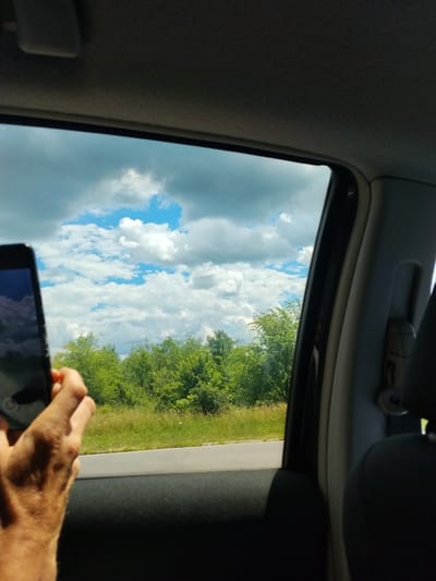 A person's hand holds a smartphone to the right of the frame, capturing a view from inside a vehicle in Budești, Moldova. The smartphone screen displays a reflection of the sky and clouds. Outside the vehicle window, a landscape unfolds with lush green trees and tall grass bordering a grey asphalt road. The sky is a vibrant blue, dotted with scattered white clouds, some of which appear to be storm clouds. The interior of the vehicle is visible in the foreground, including a dark grey headrest and part of the car door. The overall scene suggests a journey through the Moldovan countryside on a day with dynamic cloud formations.