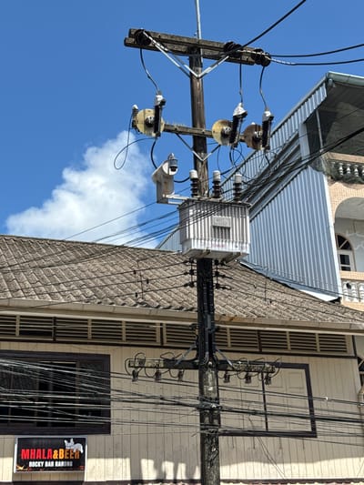The image shows a utility pole with electrical transformers and wires in the city of Ranong, Thailand. A sign at the bottom left reads "MHALL&BEER ROCKY BAR RANONG". The pole is situated in front of buildings with tiled roofs and paneled walls. The sky is blue with some white clouds. There are no people visible in the image.