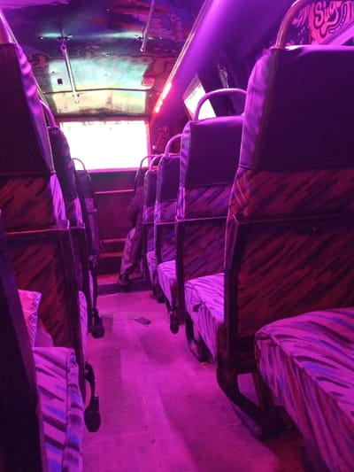 The image depicts the interior of a public transport vehicle, presumed to be a matatu, located in Kinanie ward, Kenya. The scene is dominated by a pervasive purple-magenta light emanating from linear LED strips along the ceiling and sides.

The perspective is a low-angle shot looking down a central aisle, flanked by rows of passenger seats on both sides. The seats feature dark, possibly black, upper backrests, while the lower backrests and seat cushions are upholstered in a striped fabric with patterns incorporating purple, pink, and darker tones. The seat frames are dark-colored.

One individual is partially visible, seated on the left side of the aisle, approximately midway down the vehicle. Only their lower legs and feet are discernible; they are wearing dark trousers and dark shoes.

The floor of the aisle is dark and reflects the purple illumination. A dark-colored disposable face mask is visible lying on the floor in the central aisle. The ceiling above is decorated with custom artwork or graphics and includes metal grab rails. A large, rectangular window at the far end of the aisle emits bright, diffused natural light, contrasting with the artificial interior lighting. No active interactions or specific actions are observed beyond the individual being seated.