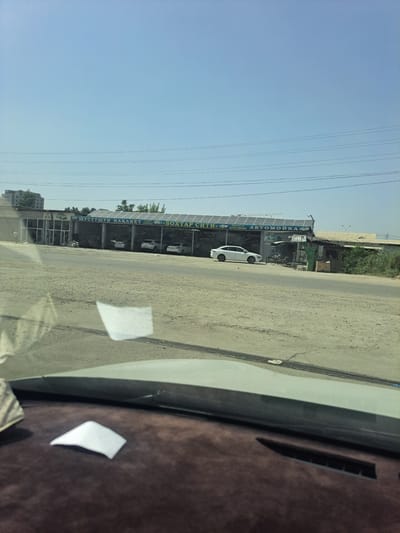 The image shows a car wash in the city of Bokhtar, Tajikistan. The car wash has a large sign with text in Cyrillic and Latin script. There are several cars parked at the car wash, and one white car is being washed. The environment is dusty and arid, with a clear blue sky and power lines overhead. The foreground shows the interior of a vehicle, including the dashboard and windshield.