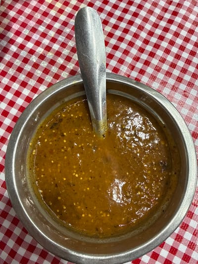 A close-up, overhead view depicts a metal bowl containing an amber-brown salsa with visible seeds and specks of darker ingredients. A silver-colored metal spoon is partially submerged vertically within the salsa, its handle extending upwards and slightly to the left. The bowl is positioned on a surface covered by a red and white checkered (gingham) tablecloth. The scene is located in the city of Nezahualcóyotl, Mexico.