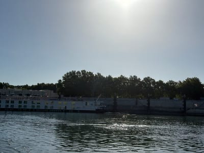A wide, low-angle shot captures a sunlit river in Arles, France, under a clear, pale blue sky. The sun is high and bright, creating a strong glare that washes out the upper portion of the image. In the mid-ground, a long, white river cruise ship is docked along a concrete embankment. The ship features multiple decks with numerous windows, some reflecting the sunlight. Behind the embankment, a dense line of dark green trees forms a backdrop. The river in the foreground is dark blue and shows small, sparkling reflections from the sun. The water appears slightly choppy, with ripples disturbing the surface. The overall scene depicts a tranquil daytime view of a waterway in Arles.