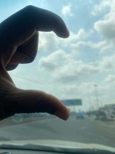 The scene presents an outdoor road environment in Owode-Elede, Nigeria. In the left foreground, a human hand is silhouetted against the bright sky, with fingers curled into a C-shape. The photograph originated from within a vehicle, indicated by a dark dashboard or interior ledge visible at the bottom. Beyond the foreground elements, a multi-lane asphalt road recedes into the distance under a sky characterized by scattered stratocumulus clouds and atmospheric haze. Multiple indistinct vehicles are present on the roadway, one appearing light blue in the mid-distance. An overhead gantry supports a large rectangular road sign, segmented in blue and green, spanning the highway. Several utility poles are distributed along the road's edges.