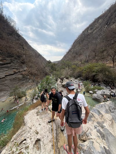 The image displays a natural canyon or valley environment near San Miguel Atlapulco, Mexico, characterized by steep, sparsely vegetated hillsides and a clear, light blue-green river flowing through the bottom. In the immediate foreground, three individuals are positioned on a large, light-colored rocky outcrop, appearing to descend towards the river below. A male on the right, viewed from behind, wears a white t-shirt, pink shorts, a grey baseball cap, and pink flip-flops, carrying a grey JanSport backpack. To his left, another male, facing partially left, wears a dark t-shirt, dark shorts, dark athletic shoes, and a dark cap, grasping a yellow rope that extends downwards. Behind him, a female, also viewed from behind, wears a dark top and dark shorts, carrying a dark backpack.
The canyon walls are composed of layered grey rock formations on the left and steeply rising hillsides covered in dry, brown vegetation on both sides, suggesting a dry season. The sky is largely overcast with prominent cloud formations. In the midground and background, numerous other individuals are visible along the riverbanks and in the water, some swimming or wading, indicating a recreational area. The overall scene depicts a group navigating a rocky descent into a natural river setting populated by other recreationists.