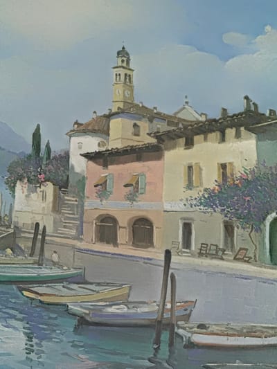 This is a painting of a canal-side village scene. In the foreground, several small boats are moored in a body of water. Behind the boats, a stone embankment separates the water from a street lined with buildings. The buildings are painted in various pastel colors, with a prominent clock tower visible in the background. Trees and flowering bushes add color to the scene. The sky is a pale blue with scattered white clouds. The overall impression is one of a peaceful, sunlit day in a European town. The scene does not depict any recognizable landmarks of Borgosatollo, Italy.