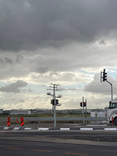 The image shows a street scene in Rosh Haayin, Israel. The weather appears cloudy and overcast. There is a road with traffic lights displaying red signals. Utility poles with power lines are visible along the roadside. Orange and black striped traffic barriers are present, and a white truck is partially visible at the edge of the frame.