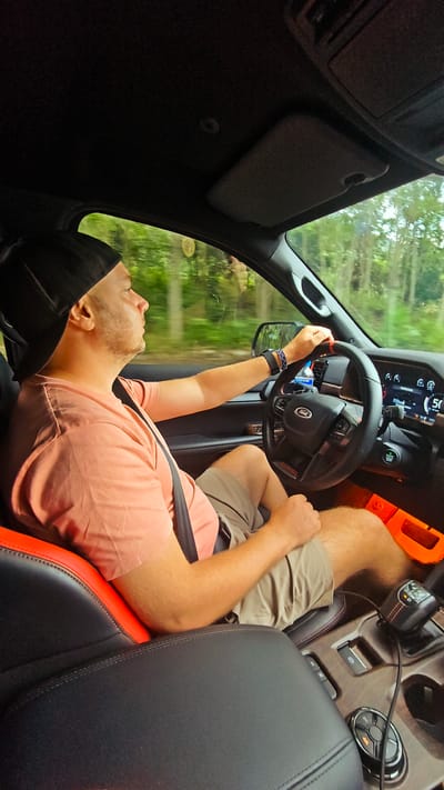 A man is driving a Ford truck. The man is wearing a black baseball cap, a salmon-colored shirt, and khaki shorts. He has a seatbelt on. The dashboard indicates the vehicle is moving at 50 kilometers per hour. Trees are visible through the window. The car's interior is black with red accents. It is difficult to ascertain if the photo was taken in Minh Tan Commune, Vietnam without corroborating visual cues.