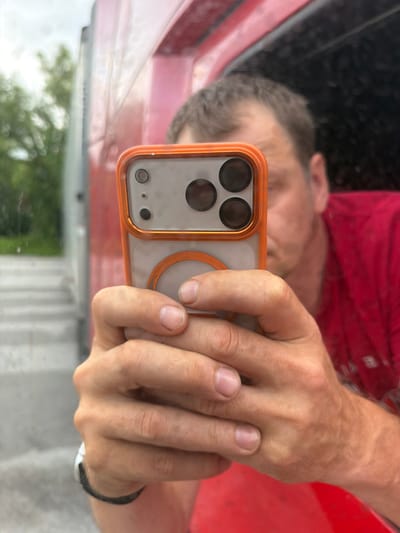 A man is holding a smartphone in his hands, taking a selfie. The phone has an orange case and a camera array with three lenses. The man is wearing a red shirt. He is likely inside or next to a red truck, as reflections of the truck's interior and exterior are visible. The background appears to be an outdoor urban environment with trees and what looks like stairs. The image is a close-up of the man's hands and phone, with his face partially obscured. The location is identified as Wildwood, United States.
