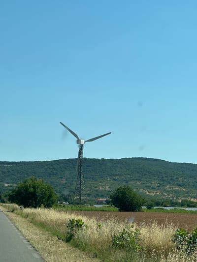 The image shows a wind turbine standing tall in a rural landscape under a clear blue sky. The turbine has a silver-colored nacelle and two large, white blades, one of which is angled upwards and to the right, and the other upwards and to the left, suggesting it is either stationary or slowly rotating. The turbine is mounted on a tall, dark-colored lattice tower.
In the foreground, there is a paved road on the left side of the image, bordered by dry, golden-colored grass and some green bushes. Beyond the road and grass, there is a field with brown soil and sparse vegetation. Further in the distance, a line of solar panels is visible, indicating a renewable energy installation.
Behind the fields and solar panels, a lush green, tree-covered hillside forms the backdrop. A few buildings, possibly houses, are scattered near the base of the hillside, some with red roofs. The overall environment is that of a sunny day in a rural area, likely in the countryside near Gorno Aleksandrovo, Bulgaria, given the context. There are no people visible in the image.